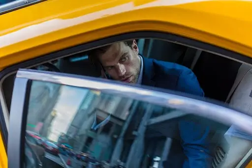 A man in a blue suit sits in the back seat of a yellow taxi, looking out the open car door at a busy city street with blurred buildings and traffic visible outside.