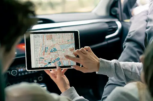 Two people sitting in a car use a tablet displaying a city map for navigation. One person points to the screen, guiding their route, while the other holds the device. Car dashboard and interior are visible.