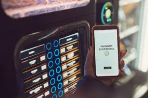 A person holds a smartphone showing a &ldquo;Payment Successful&rdquo; message with a checkmark in front of a vending machine, indicating a contactless or online payment.