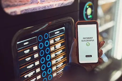 A person holds a smartphone showing a &ldquo;Payment Successful&rdquo; message with a checkmark in front of a vending machine, indicating a contactless or online payment.